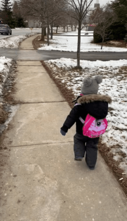 cute toddler in pom-pom hat runs down snowy sidewalk